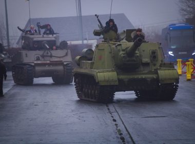Tanks drive down a road with people on top.